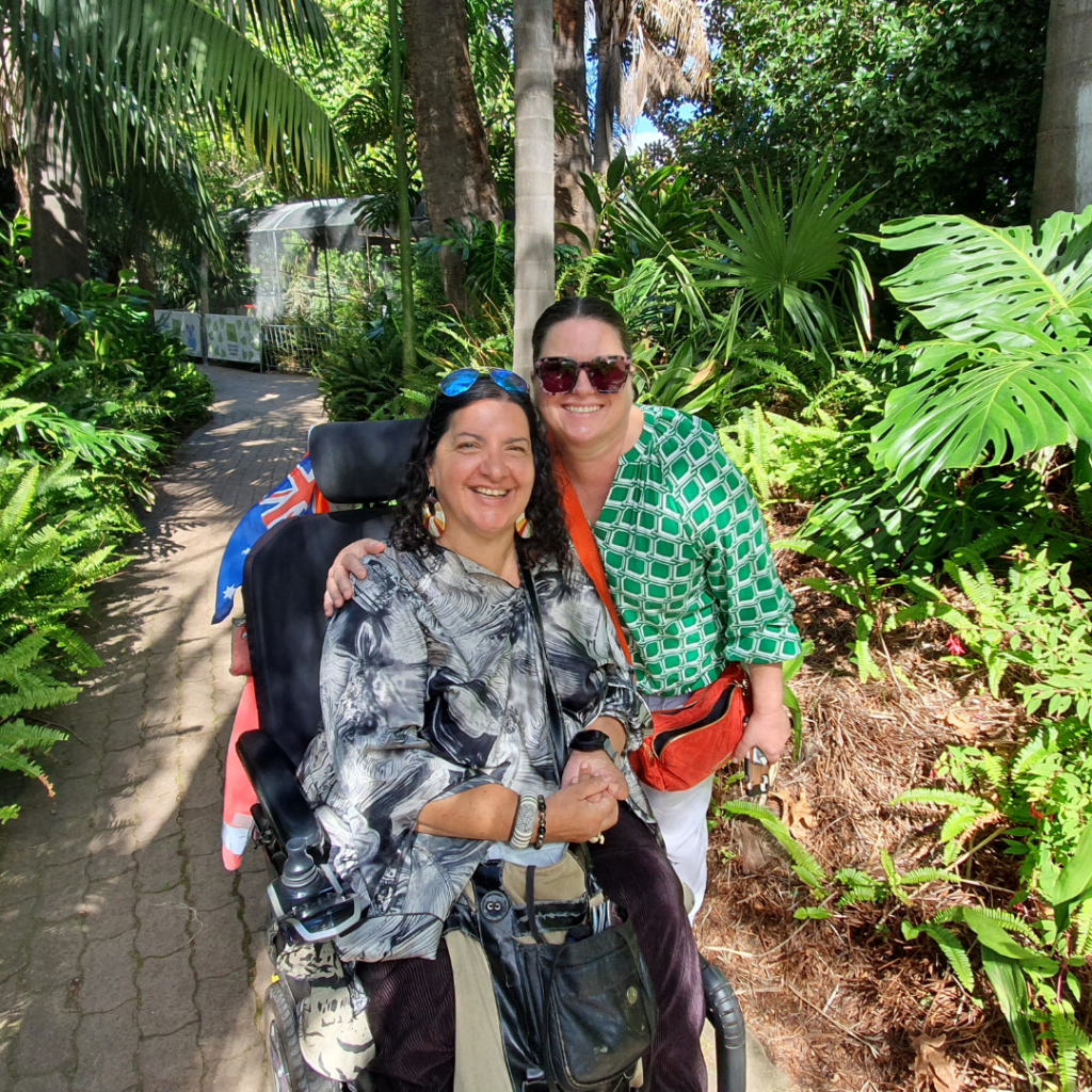Two women smiling for the camera, one in a wheelchair, surrounded by lush greenery.
