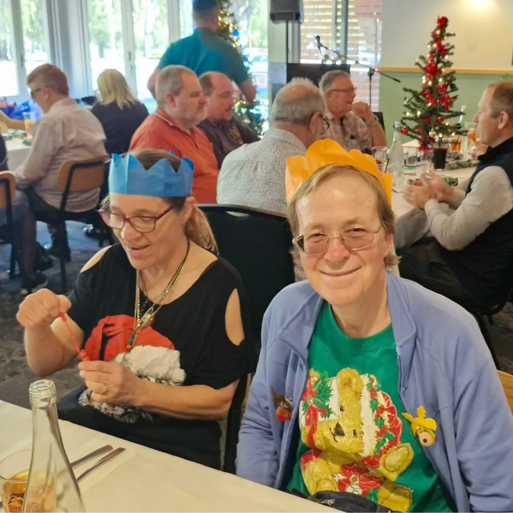 Two people wearing paper crowns sit at a holiday-themed dining table. A small Christmas tree is in the background, and other guests are seated around them.