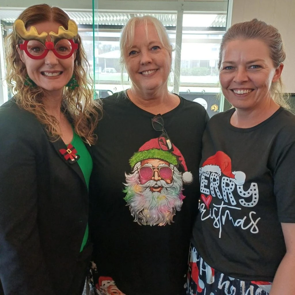 Three women smiling, two wearing Christmas-themed shirts and one with reindeer glasses.