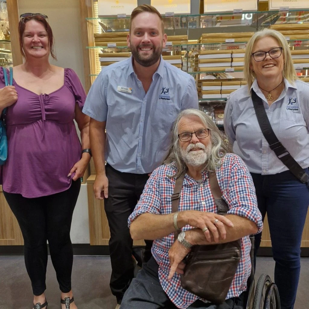 Four people posing indoors, with two standing on each side of a man sitting in a wheelchair. They are in a room with shelves in the background.