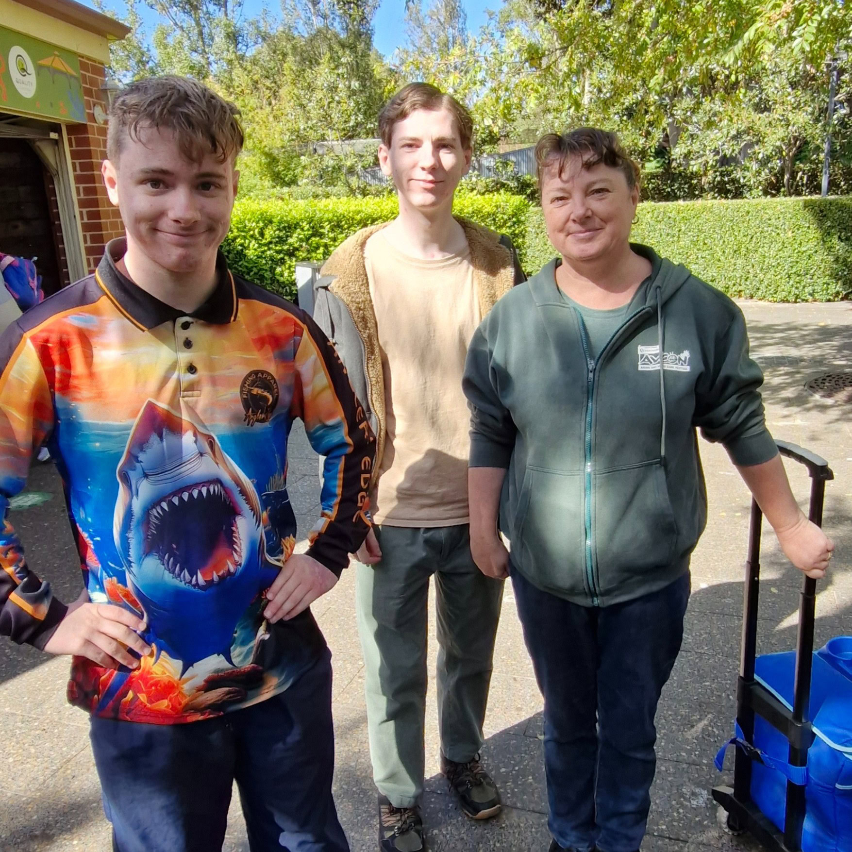 Three people stand outdoors in sunlight; one wears a colorful shark shirt, another a brown jacket, and the third a green zip-up hoodie while holding a suitcase handle.