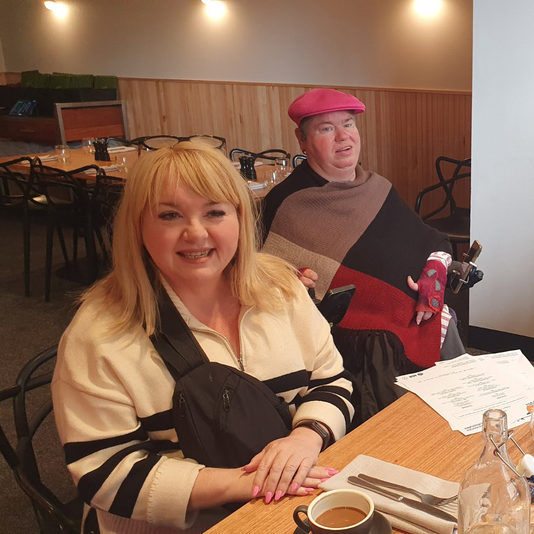 Two people are seated at a wooden table in a restaurant. The person in front has blonde hair and a striped top. The person behind wears a pink beret and a colorful poncho. A menu and coffee mug are on the table.