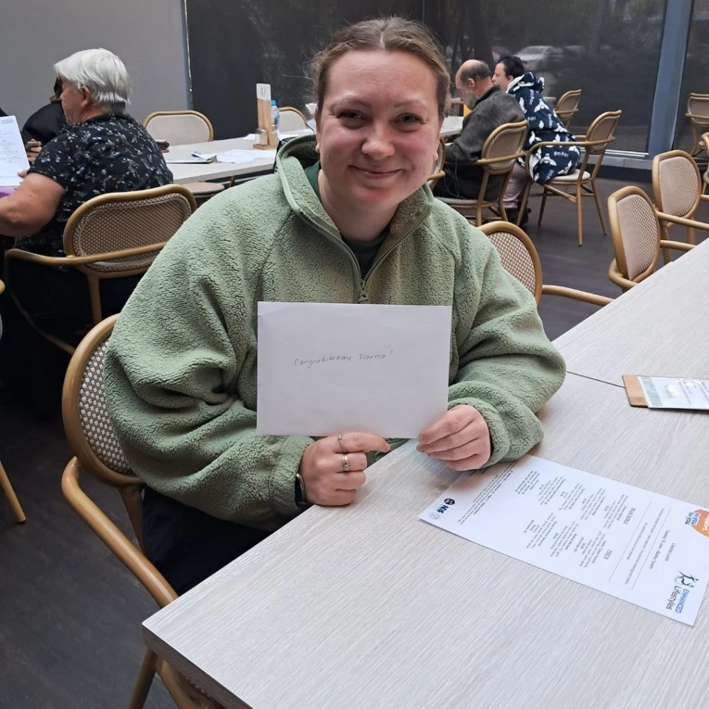 A person sits at a table holding an envelope with "Congratulations" written on it, smiling. Other people are seated and engaging in activities in the background.