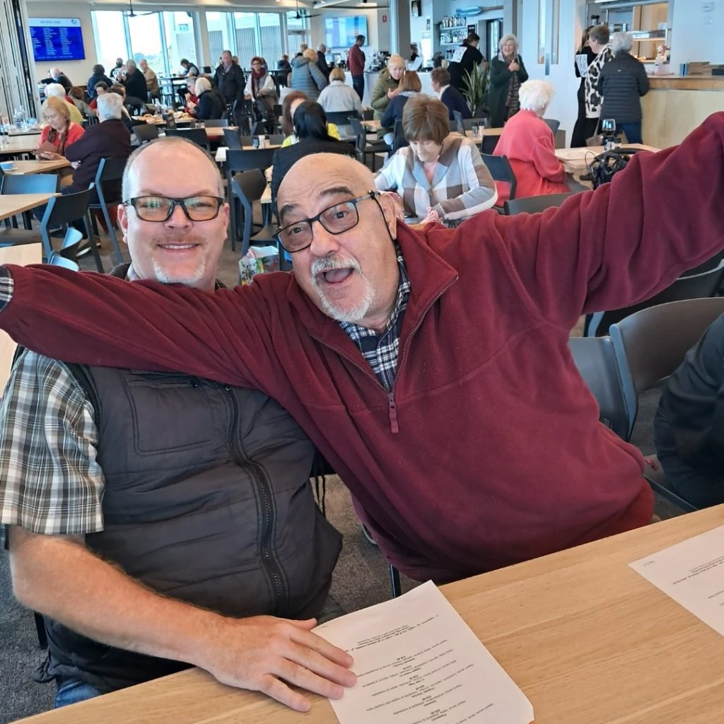 Two men are seated at a table in a crowded dining area. One man is smiling, while the other is extending his arms joyfully. Papers are on the table in front of them.