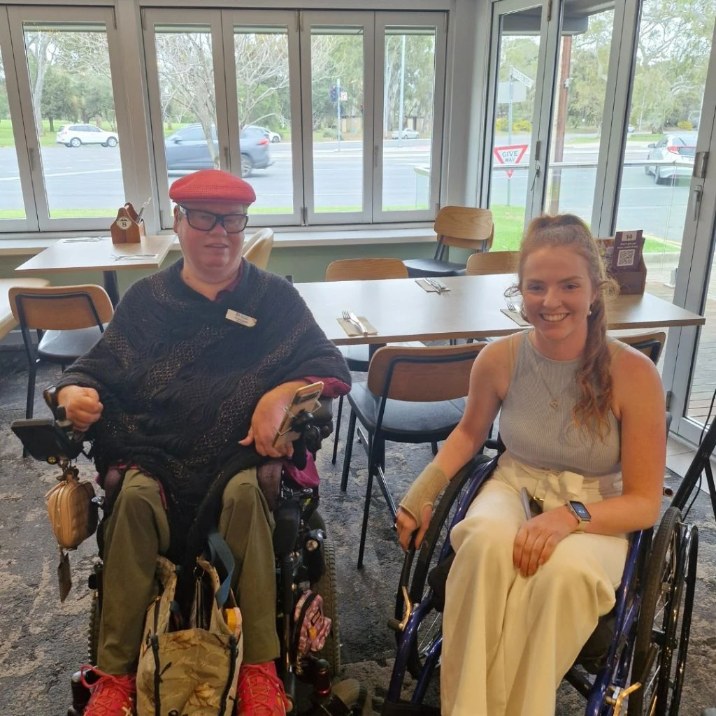 Two people in wheelchairs are sitting next to each other in a dining area. One person is wearing a red hat, glasses, and a dark poncho. The other person is wearing a light-colored sleeveless top and white pants.