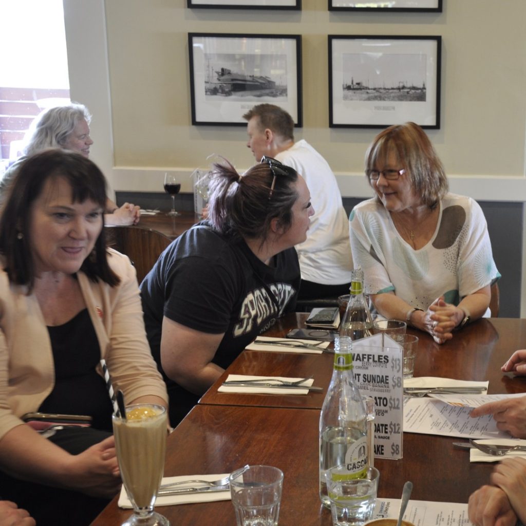 A group of people are seated around a table in a restaurant, engaged in conversation. Menus, drinks, and a bottle of water are on the table. Framed black-and-white photographs are on the wall in the background.