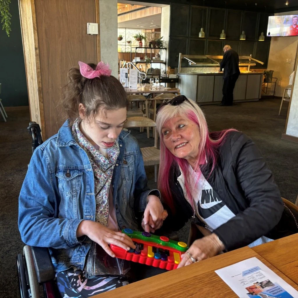 A young girl plays with a colorful toy while sitting next to a woman with pink-tinted hair in a restaurant. The woman smiles at the camera, and the girl appears focused on the toy.