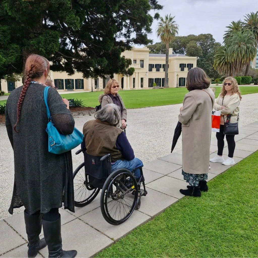 A group of five people, including an individual in a wheelchair, are gathered outside near a large building surrounded by trees and lawn.