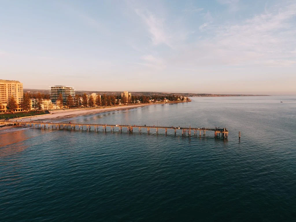 Aerial view of a coastal city with a long pier extending into the ocean, lined with buildings and a sandy beach under a clear sky.