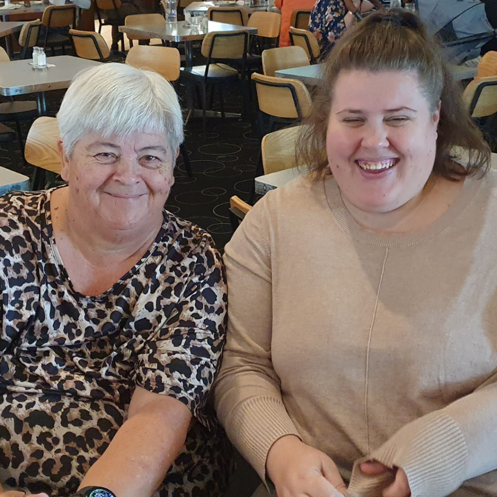 Two smiling women sitting side by side indoors, with a restaurant setting in the background.