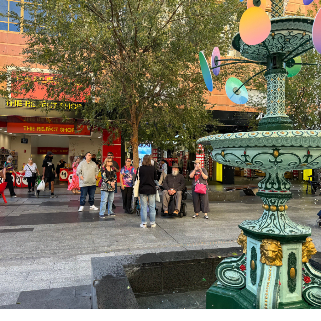 Pedestrians gathered around a colorful ornamental fountain in an urban plaza with trees and shops in the background.
