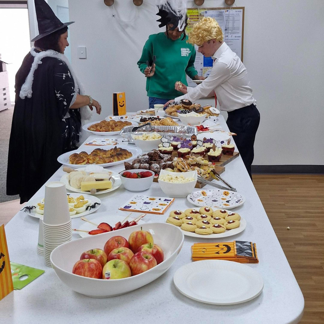 People in costumes stand by a table with Halloween-themed food, including apples, cookies, and pastries.