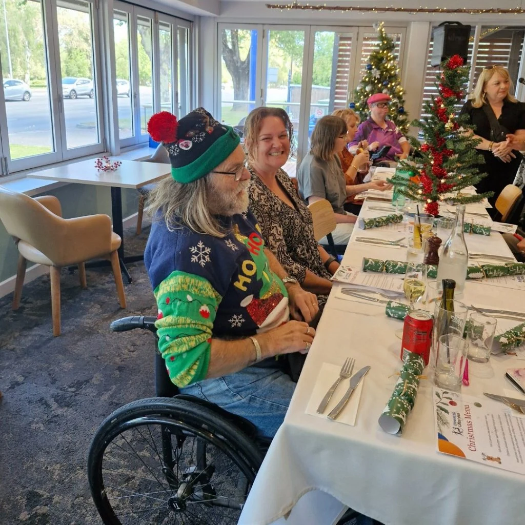 A group of people sit around a festive table with Christmas decorations, including small trees and crackers. One person in a wheelchair wears a holiday-themed sweater and hat.