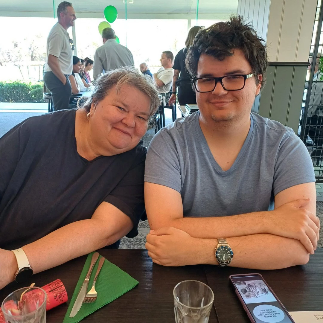 Two people sitting at a table in a restaurant, smiling at the camera. The table has drinks and table settings. Other diners and a green balloon are in the background.