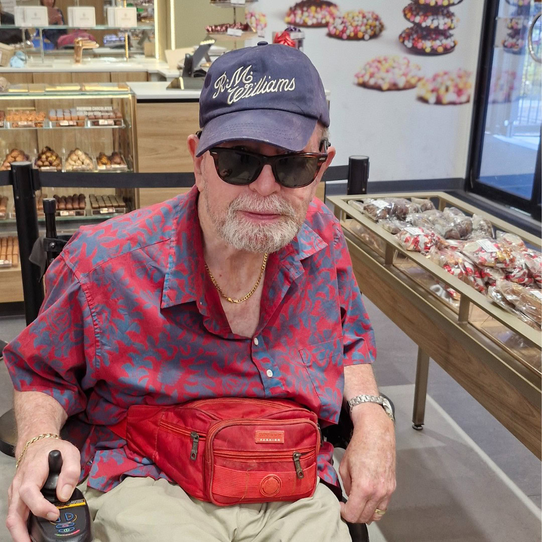 An elderly man in a red floral shirt, sunglasses, and a cap sits indoors. He holds a drink can and has a red waist bag. Baked goods are displayed in the background.