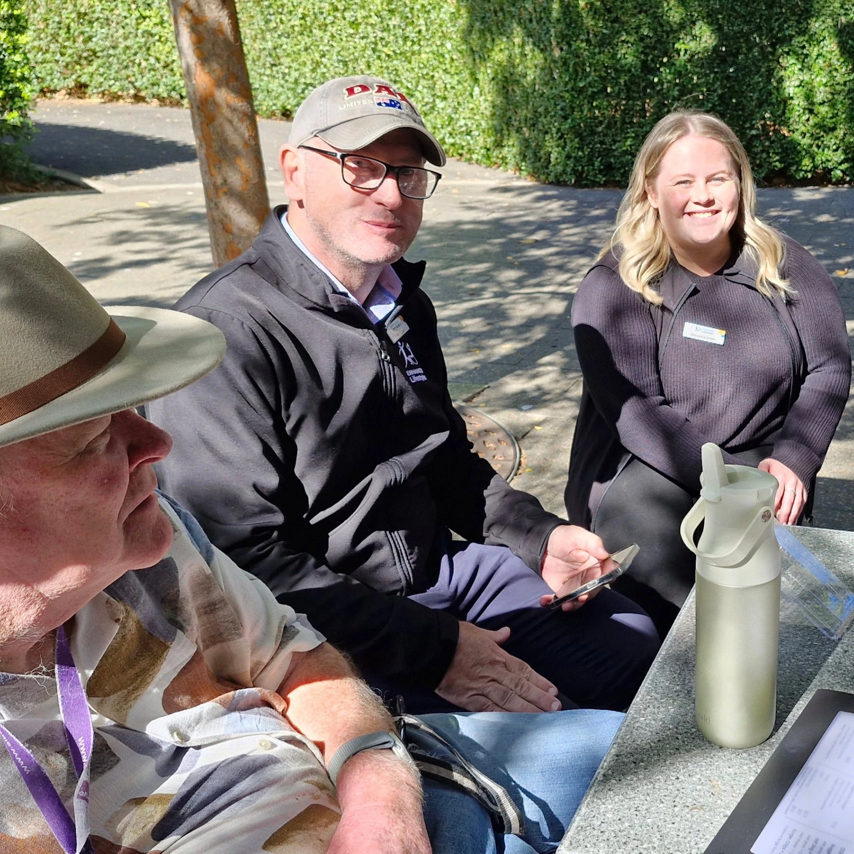 Three people sit outdoors at a table in sunlight. One holds a phone, one has a water bottle nearby, and all are dressed casually. A hedge and pathway are visible in the background.