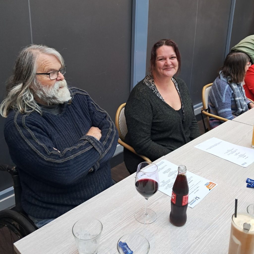 Two people sit at a table in a restaurant. The man on the left with gray hair and beard has arms crossed, and the woman on the right smiles. There are drinks, glasses, and menus on the table.