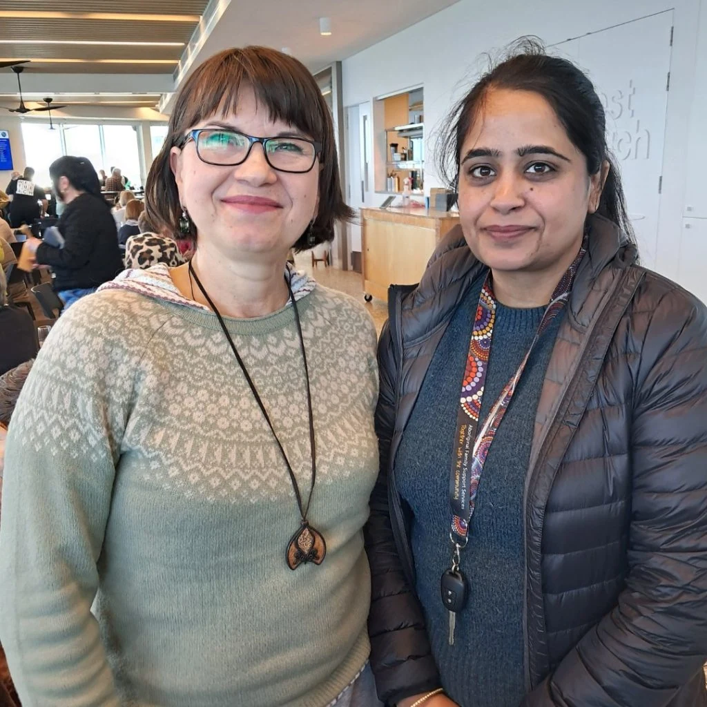 Two women stand indoors, facing the camera. One wears a green sweater and glasses, and the other wears a dark jacket and colorful necklace. A cafe setting with people and tables is visible in the background.