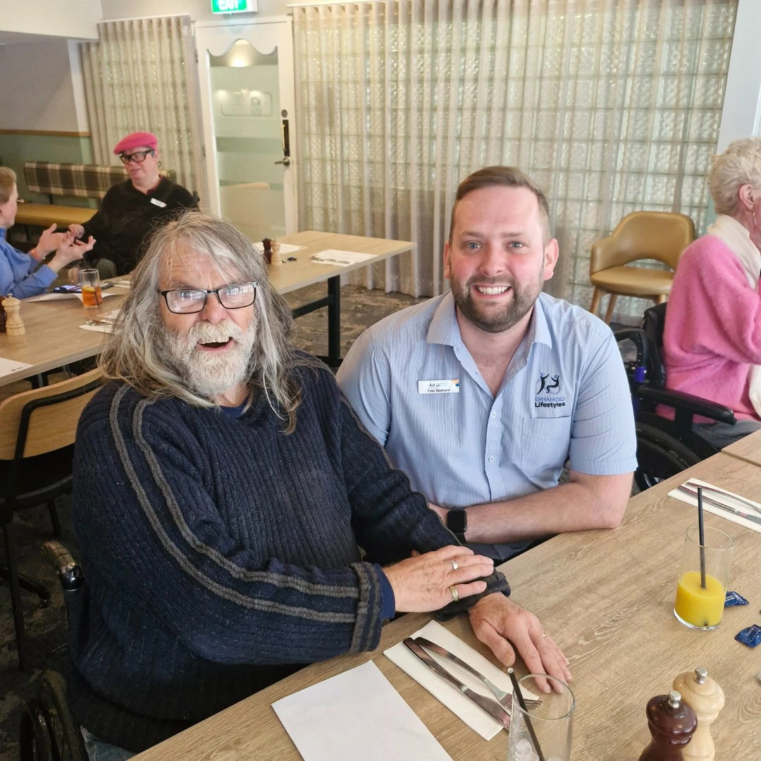 An elderly man with long gray hair and a beard sits at a table with a younger man in a blue shirt, both smiling at the camera. Other people are visible in the background inside a dining room.