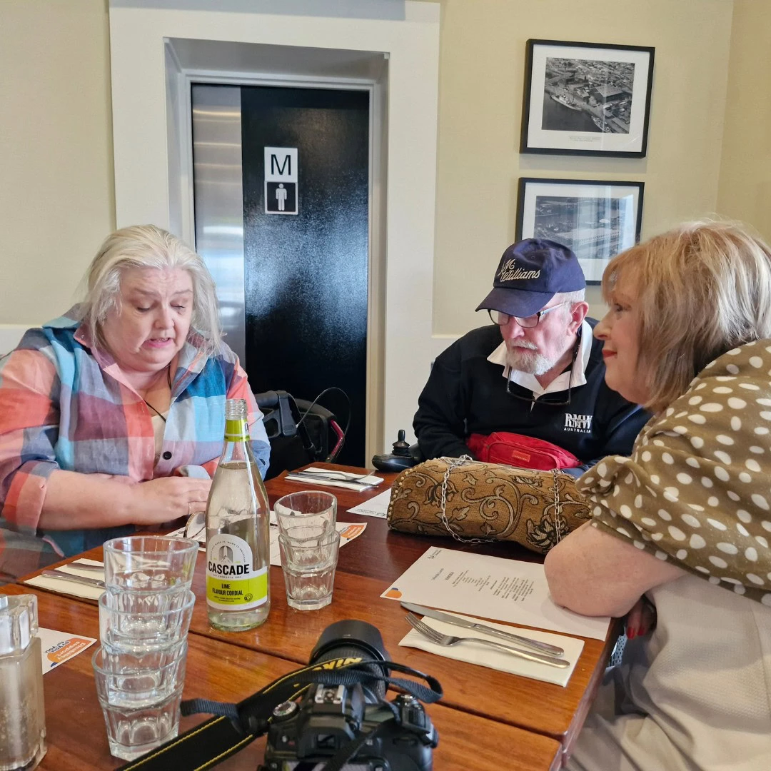 Three people sit at a wooden table in a restaurant. One woman appears to be talking while another woman and a man listen. The table has glasses, a water bottle, and a menu. A camera is visible on the table.