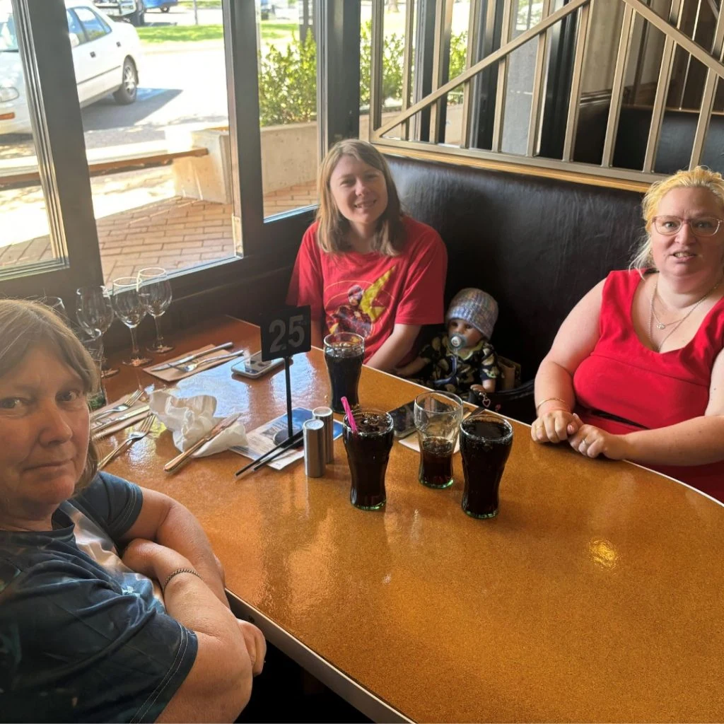 Three people sitting at a restaurant booth with drinks and a table number 25. They are inside, near a window with a view of parked cars outside.