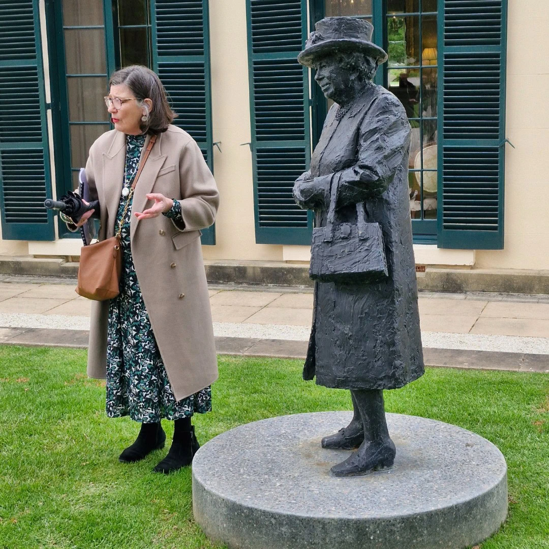 A woman stands next to a statue of a figure in a coat and hat, both on a lawn in front of a building with shuttered windows.