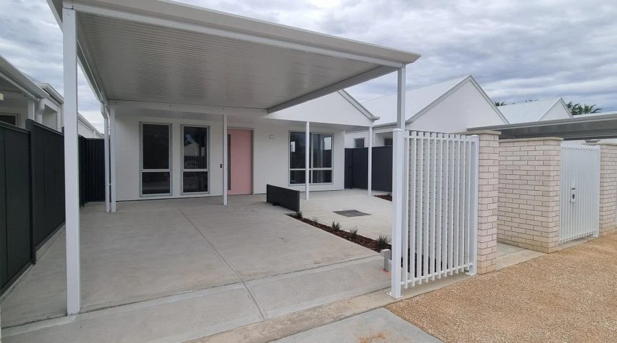 Modern white house with a covered carport, gated entrance, and small front yard under a cloudy sky.