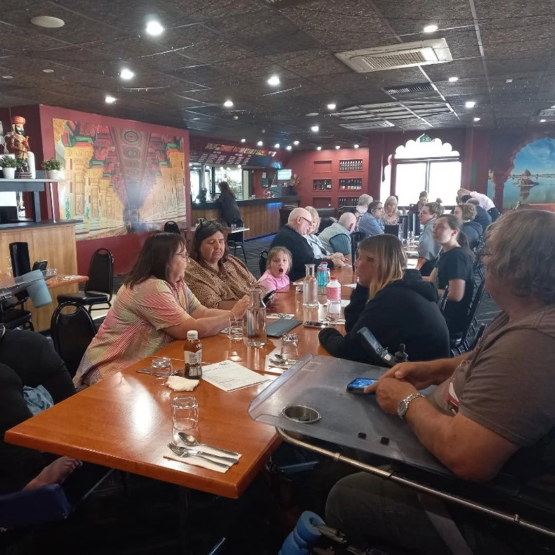 A group of people sitting at a long table in a restaurant, engaging in conversation and enjoying their meals. The background features vibrant wall art and a bar area.