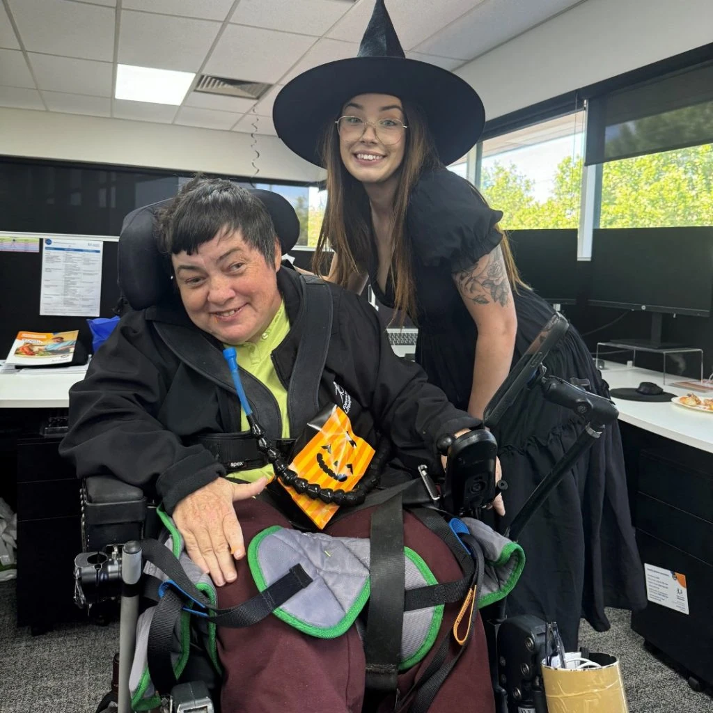 Two people smiling in an office. One is in a wheelchair with Halloween decorations, and the other is wearing a witch hat and black dress.