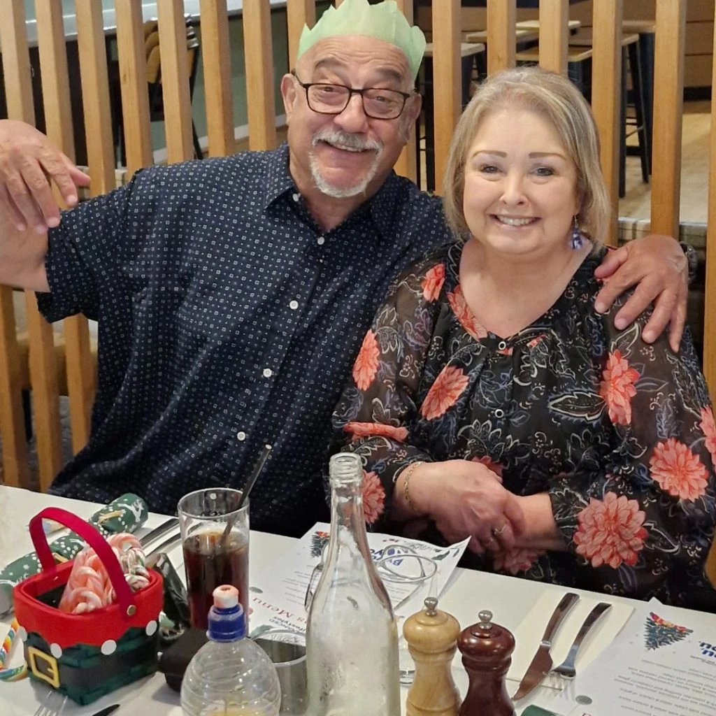 A man and woman are seated at a dining table, smiling. The table has drinks, a Christmas-themed basket, and condiment bottles. The man wears a green paper crown.