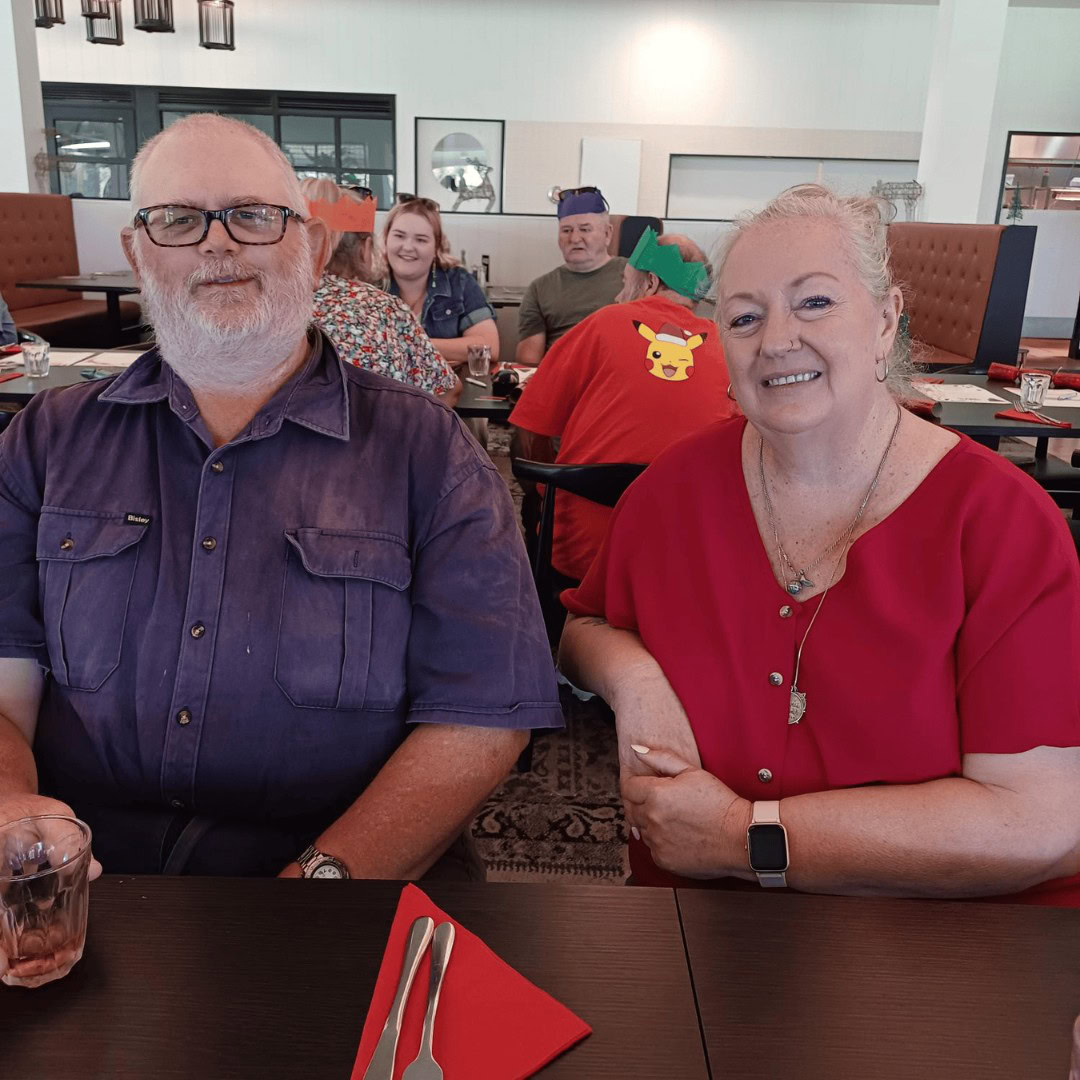 A man and woman sit together at a restaurant table with people in the background. They are smiling and positioned in the foreground.