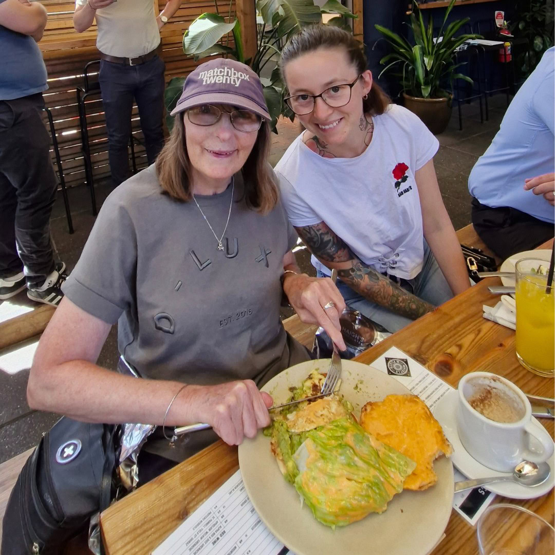 Two women sitting at an outdoor table enjoying a meal. One woman is cutting into a dish. They seem relaxed and smiling. Drinks are on the table, including a cup of coffee and a glass of juice.