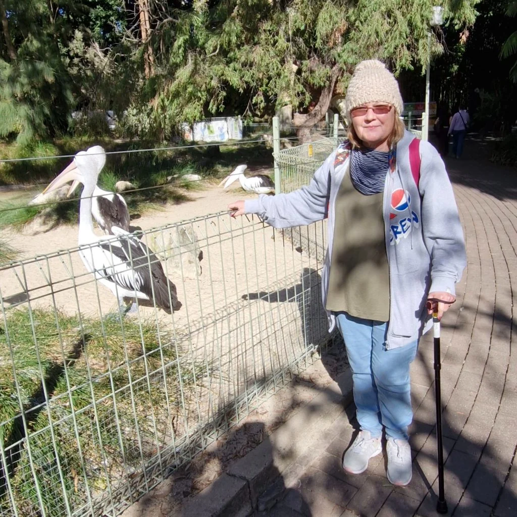 A woman stands next to a fenced enclosure with pelicans inside, holding a walking stick and wearing sunglasses, a beanie, and a gray jacket with a Pepsi logo.