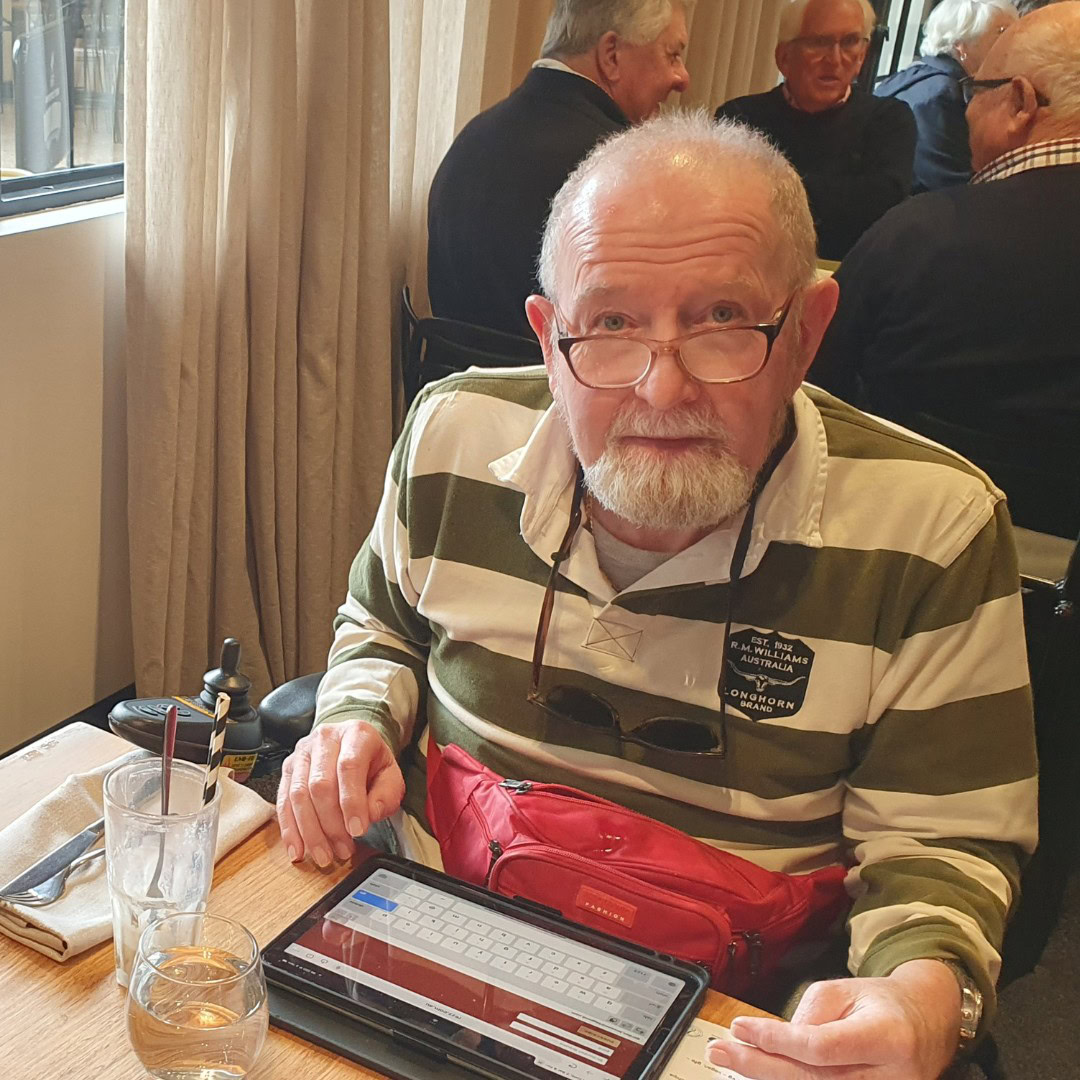 Elderly man with glasses and a striped shirt is seated at a table in a restaurant, using a tablet. Other diners can be seen in the background. A glass of water and cutlery are on the table.
