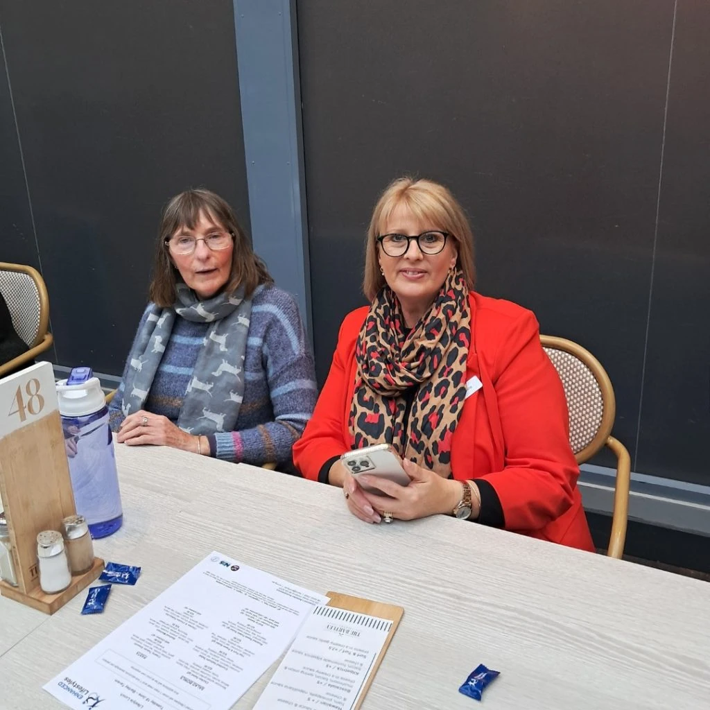 Two women seated at a table, one wearing a blue sweater and scarf, the other in a red blazer with a leopard print scarf. A menu and some packets are on the table.