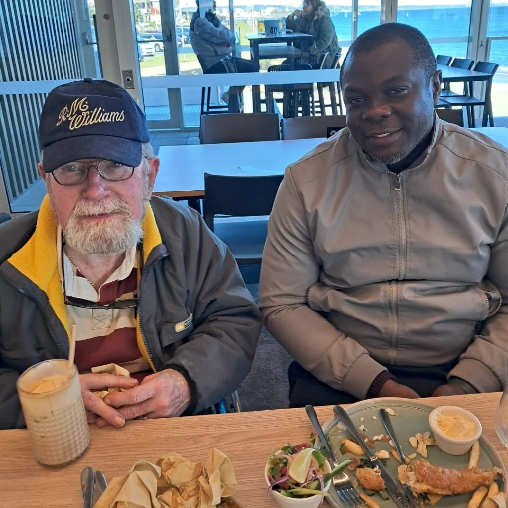 Two men sitting at a dining table with dishes of unfinished food and drinks. One man wears a cap and glasses, while the other wears a light-colored jacket. They appear to be inside a restaurant with large windows.