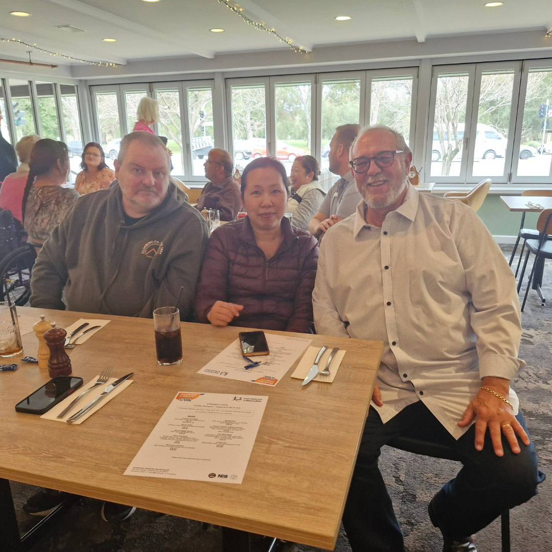 Three people sit at a table in a restaurant, with menus and utensils in front of them. The restaurant has large windows showing a view of trees outside. Other patrons are visible in the background.