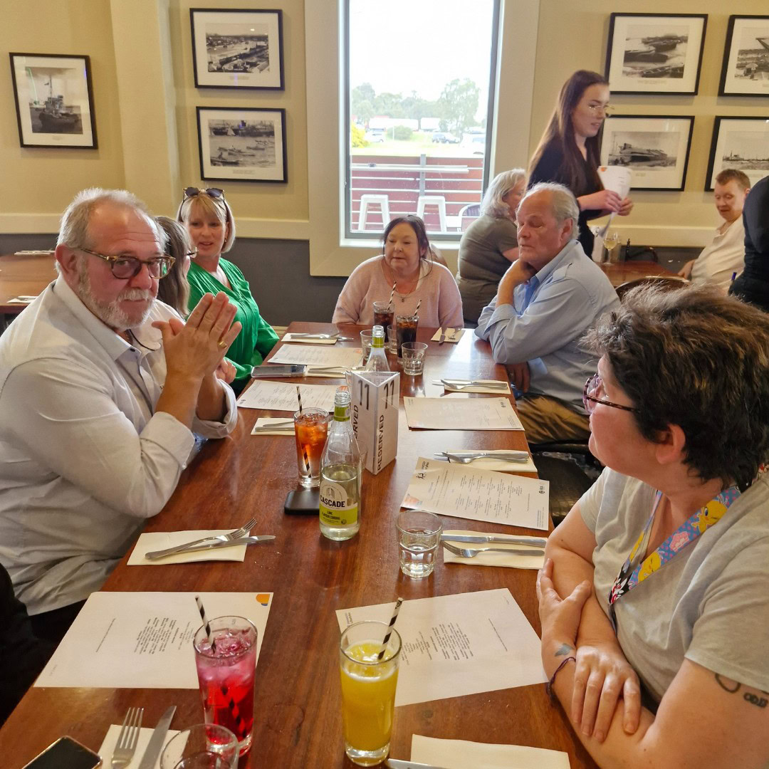 A group of people seated around a table at a restaurant, looking at menus. Drinks are on the table, and framed pictures are on the wall behind them.