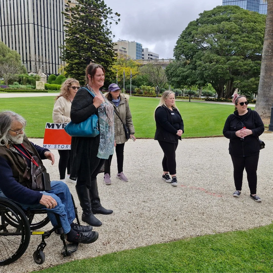 A group of people, including one in a wheelchair, stand on a gravel path in a park with trees and buildings in the background.