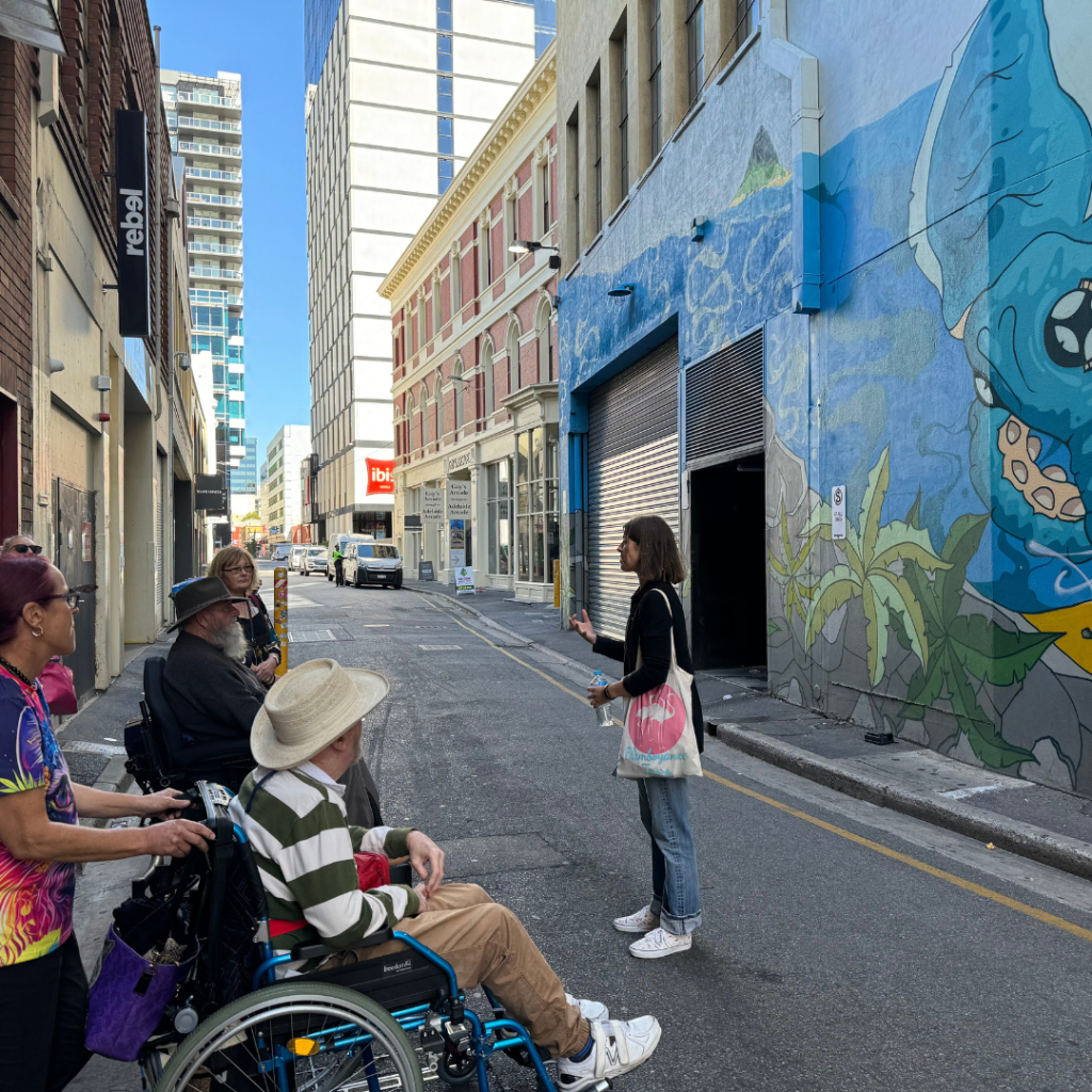 A group of people, one in a wheelchair, engaged in conversation on a sunlit street with graffiti art on the walls.