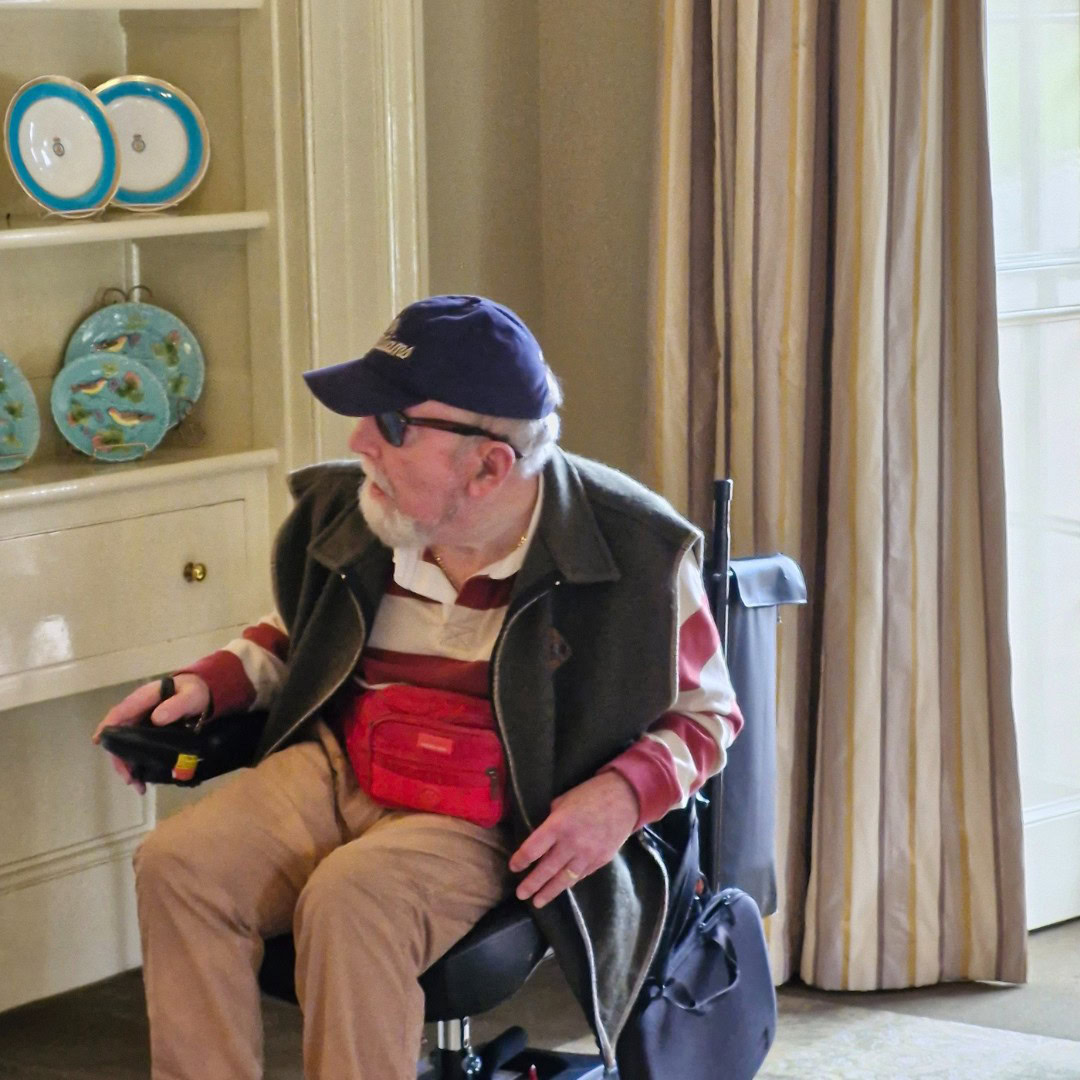 An elderly man wearing a cap and glasses sits in a wheelchair near a shelf displaying decorative plates. He holds a device and wears a red pouch bag.