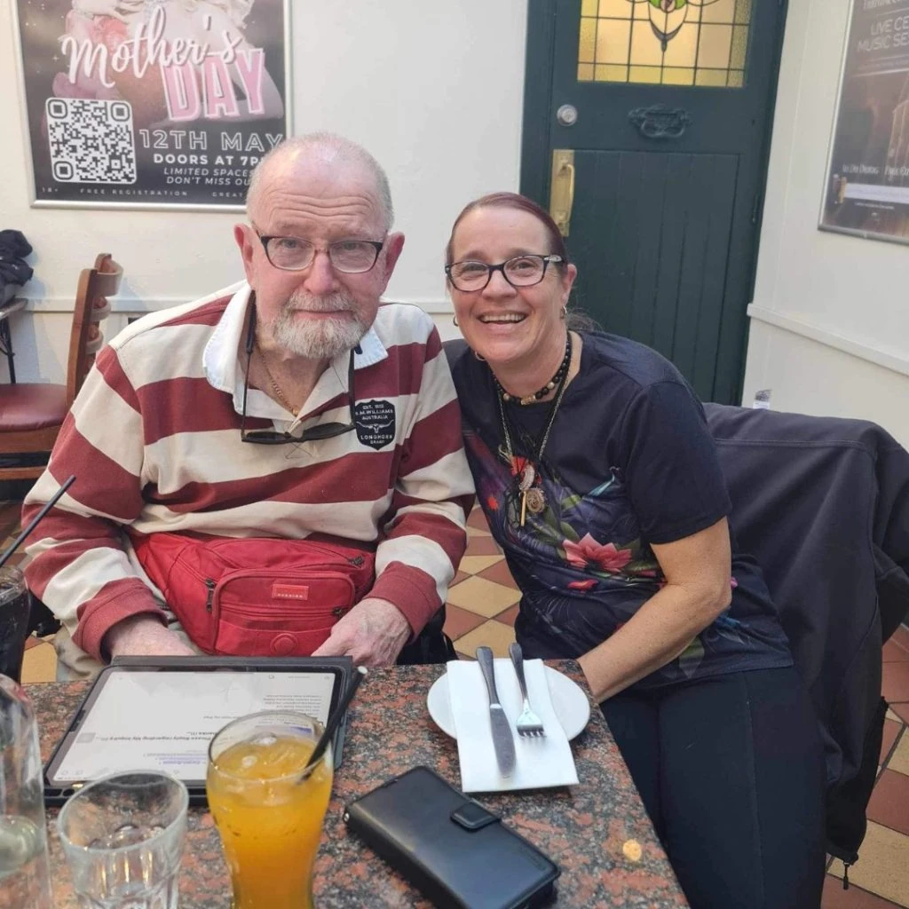 An elderly man and a middle-aged woman smiling at a restaurant table with drinks and cutlery.
