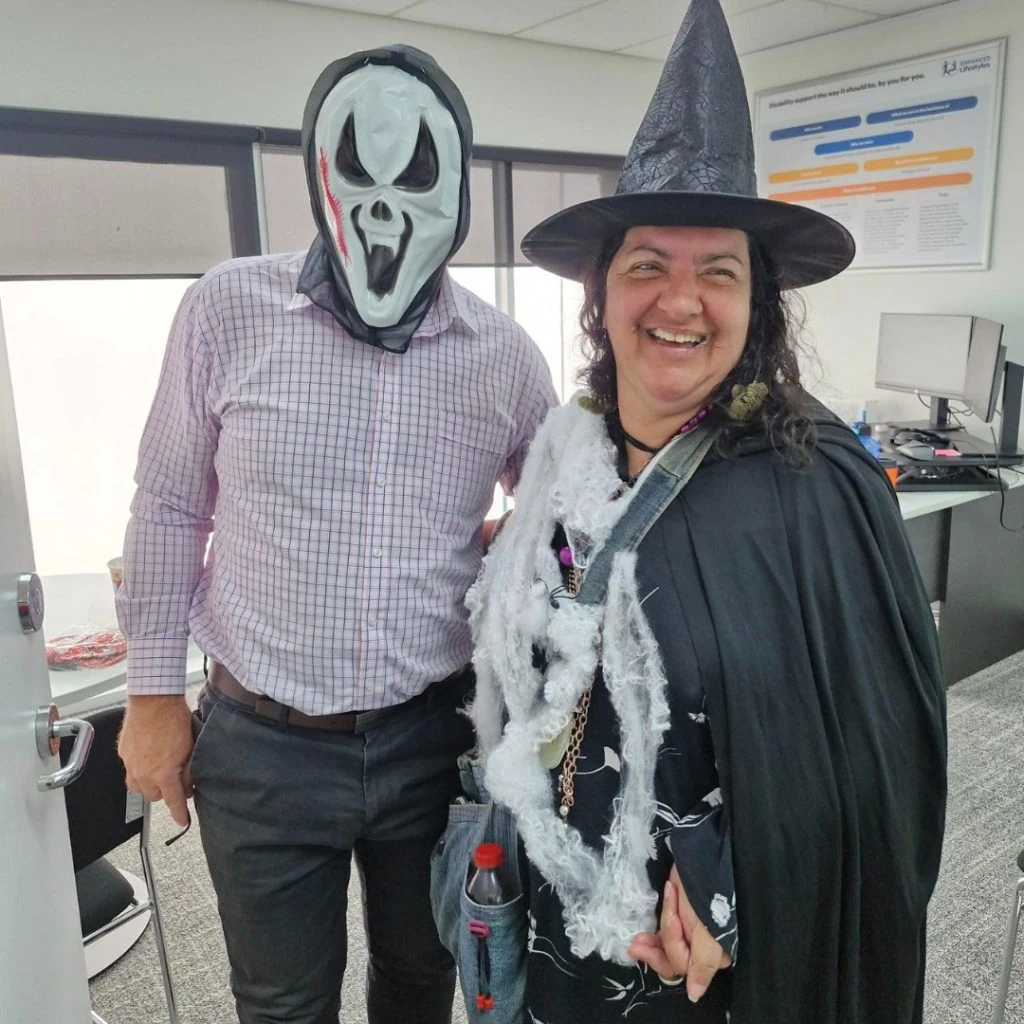 Two people dressed in Halloween costumes, one wearing a ghost mask and the other in a witch outfit, smile in an office setting.