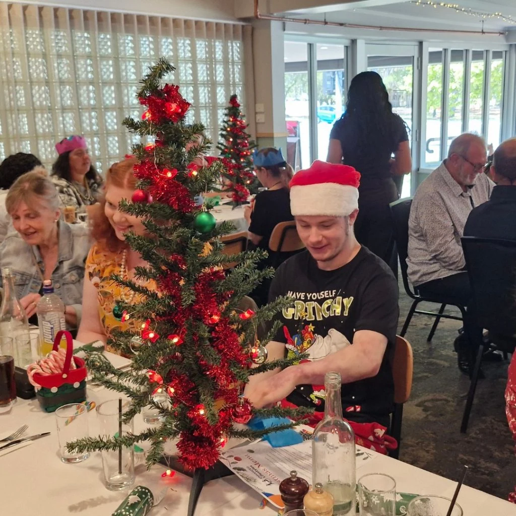 People sitting at tables decorated for Christmas, with a person in a Santa hat next to a small Christmas tree.