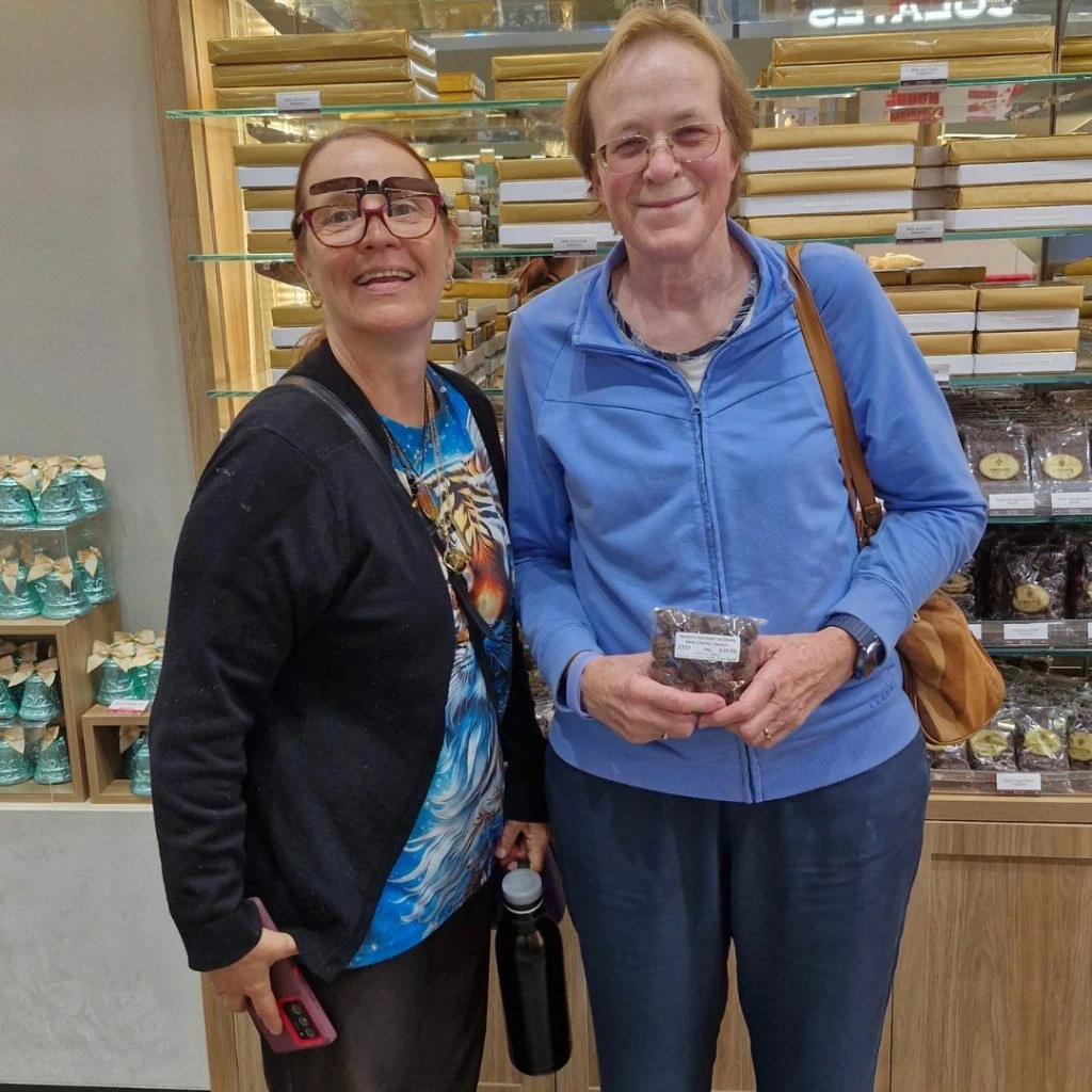 Two people standing in a bakery; one holds a pastry box. Shelves with bakery items are visible in the background.