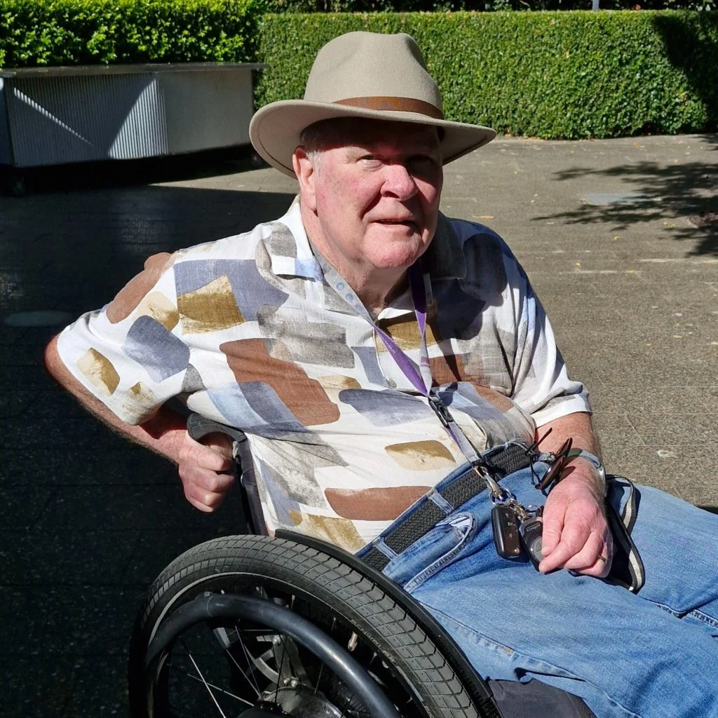An older man in a patterned shirt and hat sits in a wheelchair outdoors on a sunny day, with greenery and pavement in the background.