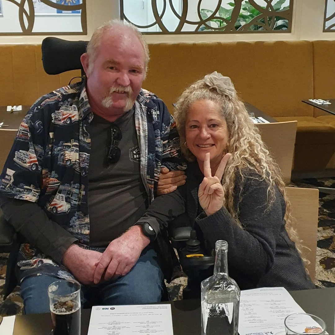 Two people sitting at a restaurant table, smiling. The person on the left is wearing a patterned shirt, and the person on the right is making a peace sign with their fingers.