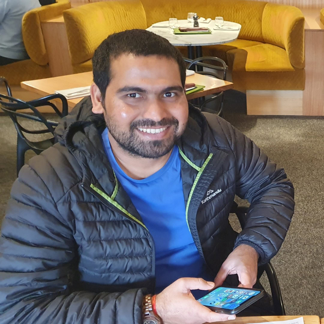 A man with a beard, wearing a black jacket and blue shirt, is sitting in a restaurant and smiling at the camera while holding a smartphone.