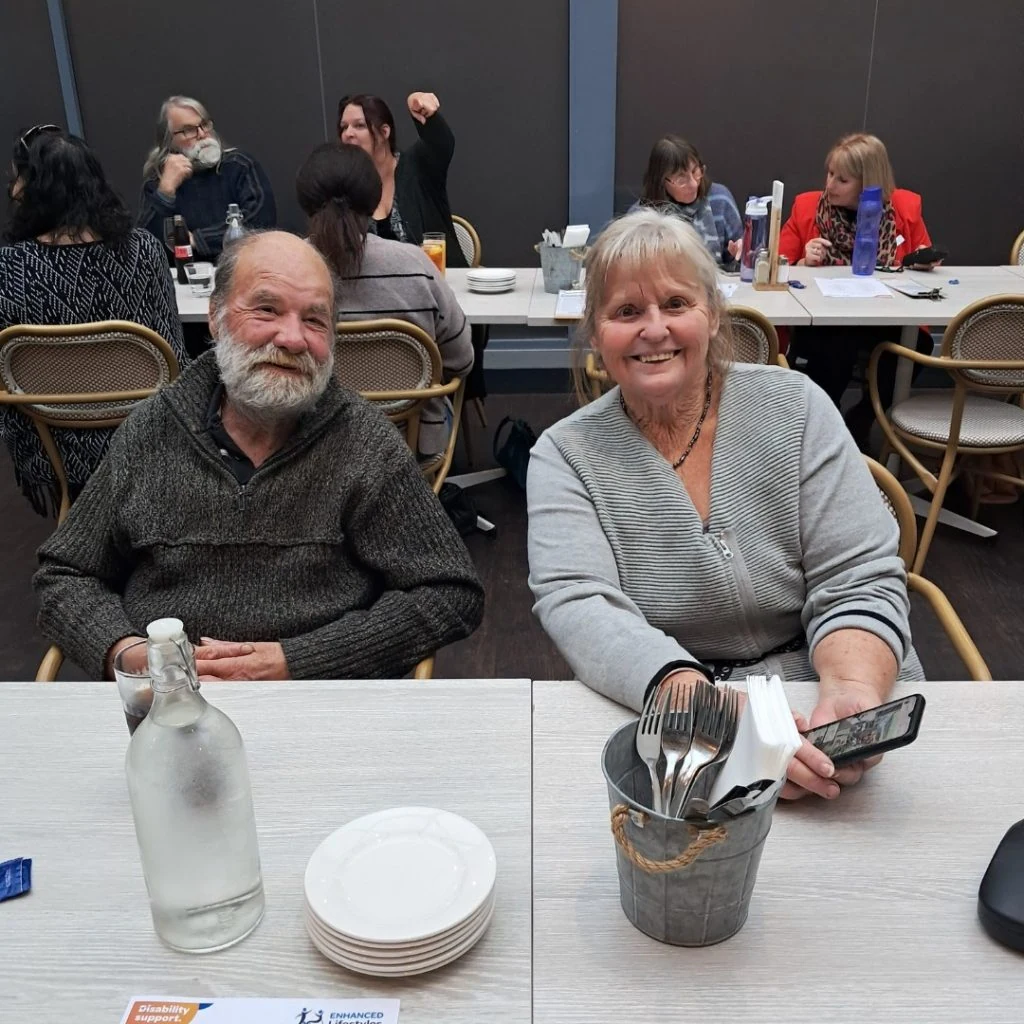 Two elderly people sit at a table in a restaurant, smiling at the camera. Other diners are seen in the background, engaged in conversation.