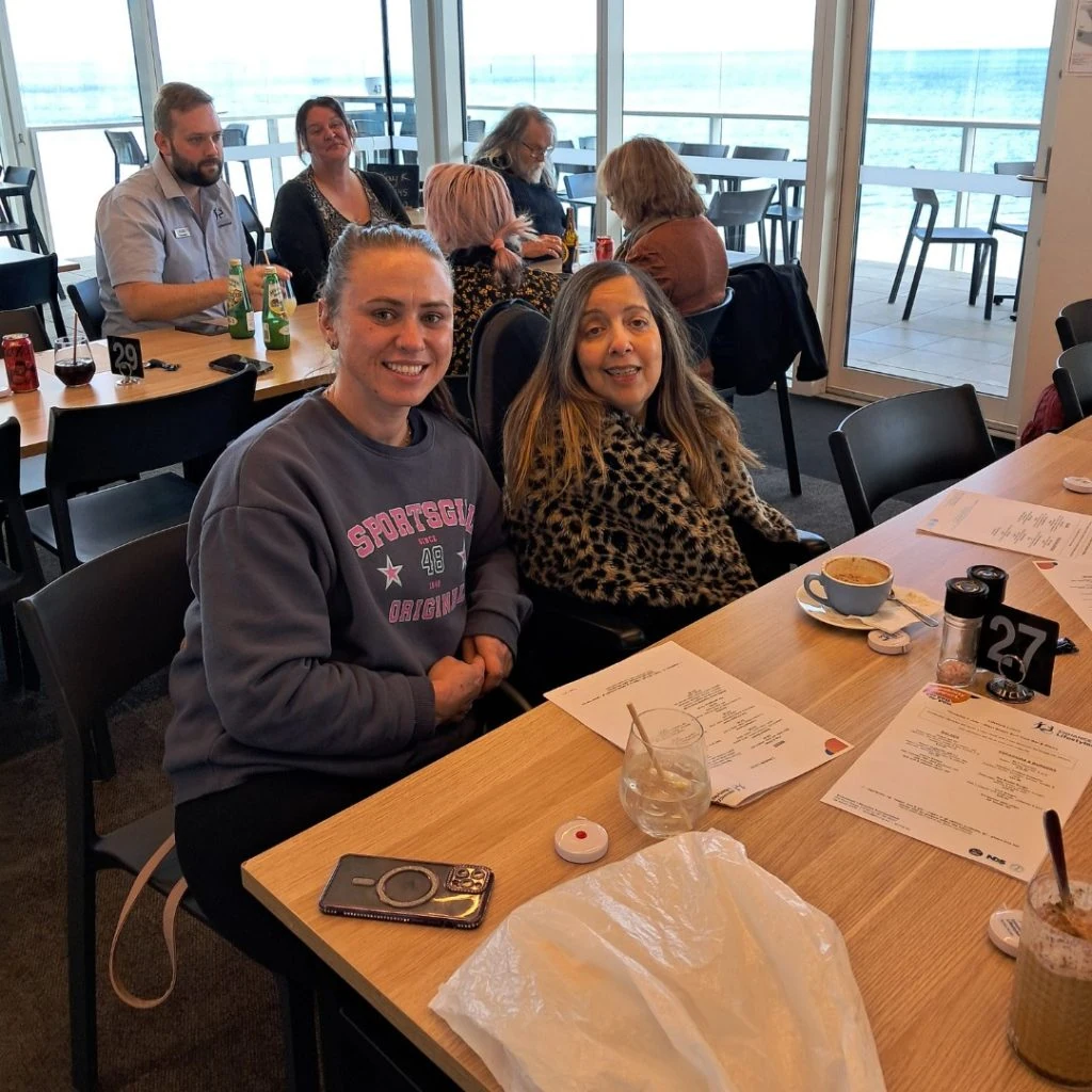 Two women seated at a table in a restaurant, smiling at the camera. Other patrons are seen in the background. The table has menus, drinks, and food items. Large windows reveal an ocean view.